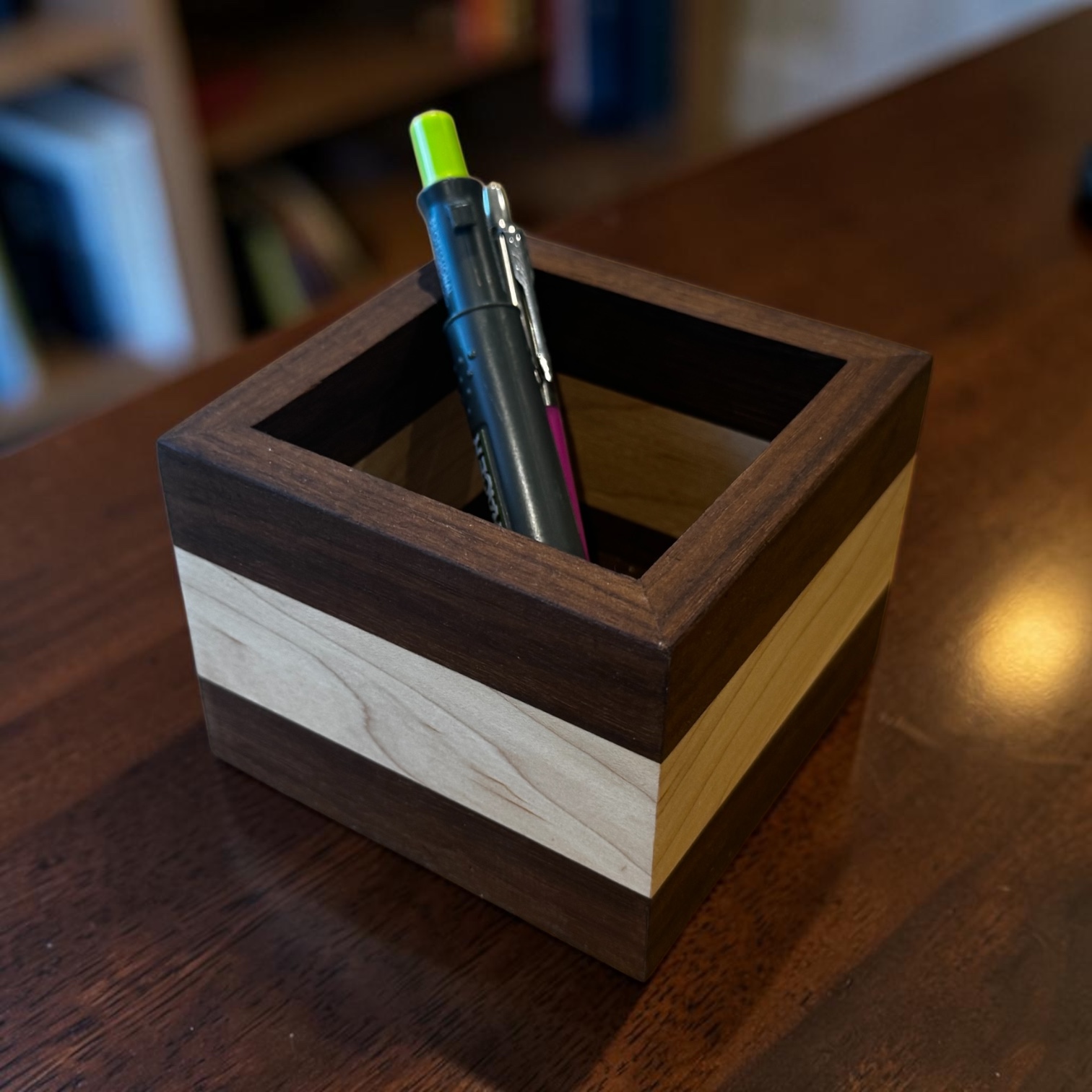 A walnut and maple square desk tidy showing pens on a wooden desk.