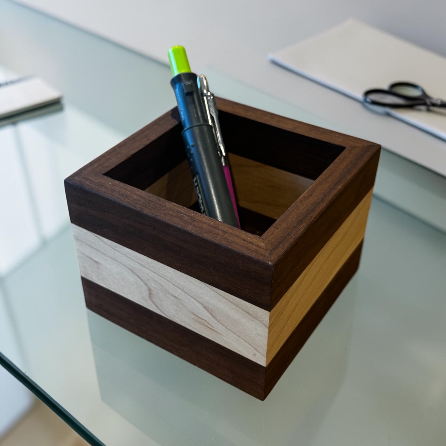 A walnut and maple square desk tidy showing pens on a glass desk.