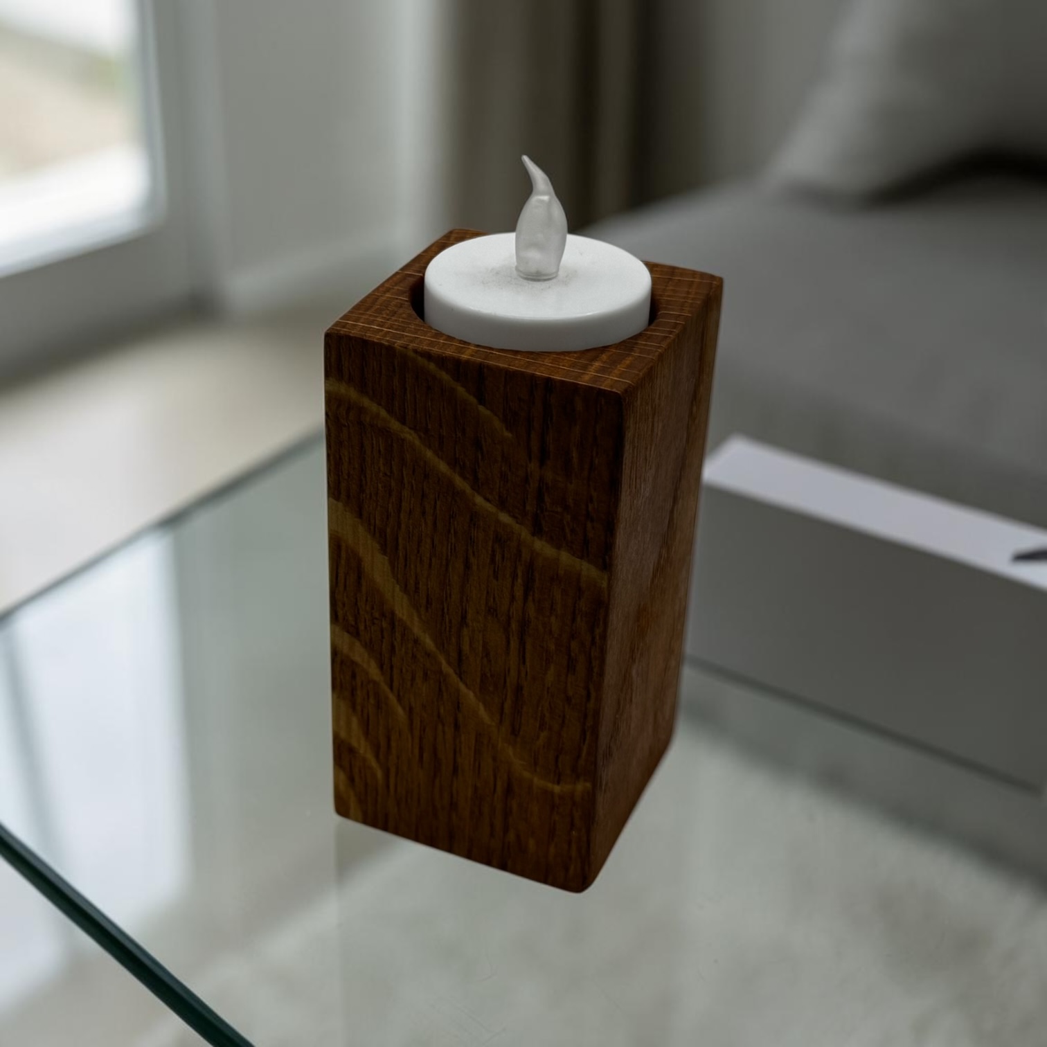 A medium tea light holder that is tall and square with an electric tea light in the top on a glass table.